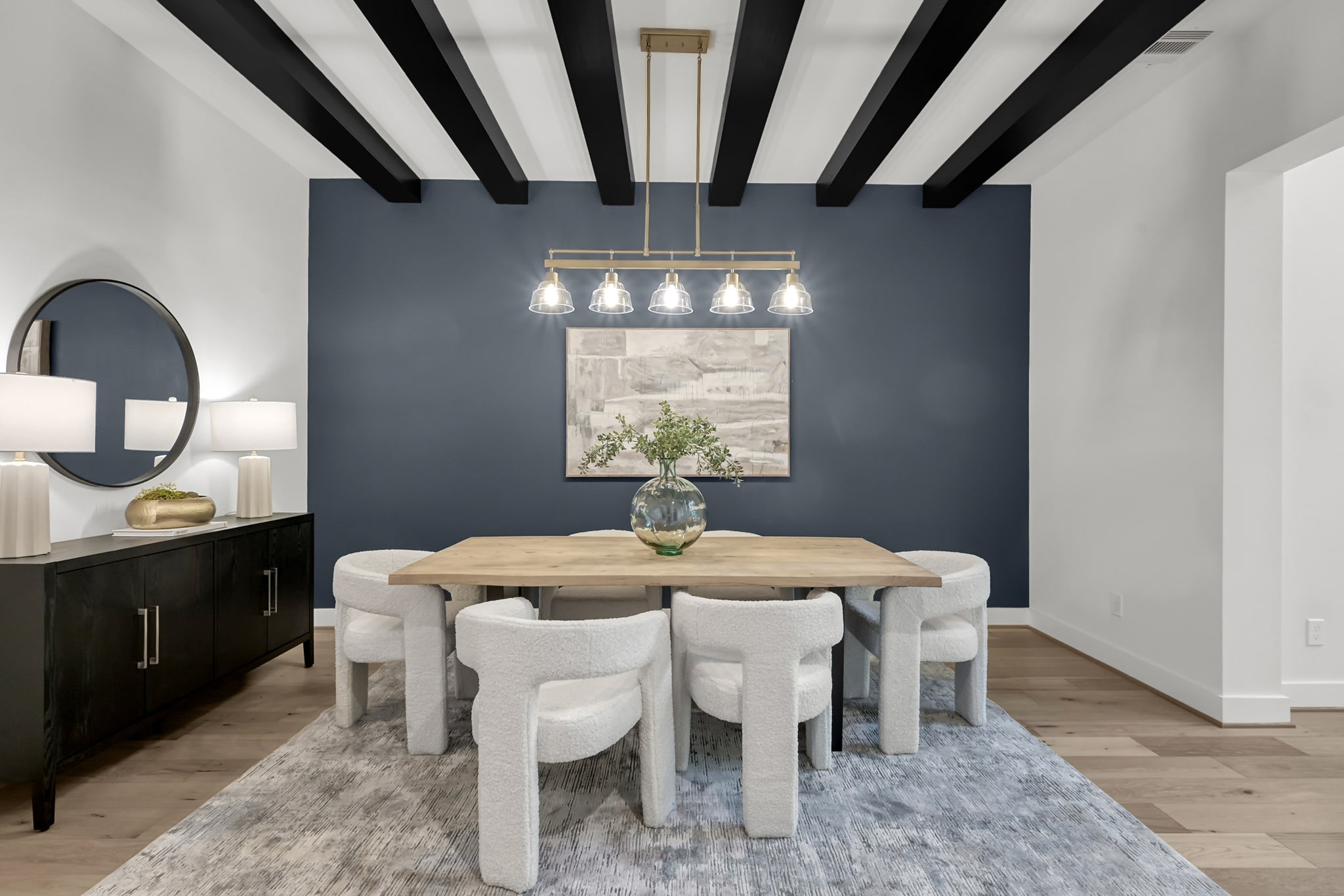 Modern dining room with a wooden table, six white upholstered chairs, a blue accent wall, black ceiling beams, a large sideboard with lamps, and a round mirror. A vase with greenery decorates the table.