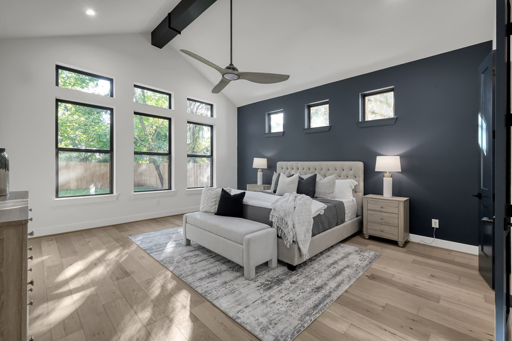 Modern bedroom with a dark accent wall, large windows, light wood floors, a tufted bed, gray and white bedding, a bench, two nightstands with lamps, and a ceiling fan. Natural light fills the room.