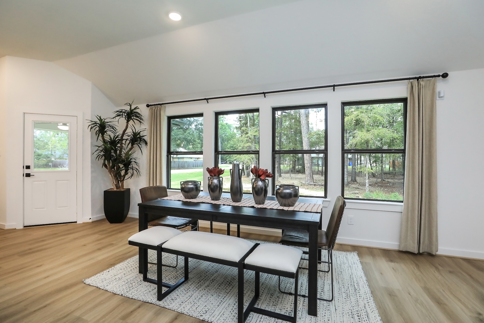 Modern dining room with a dark wood table, white cushioned benches, and three black vases with red flowers. Large windows overlook green trees, and light wood floors and a potted plant complete the space.
