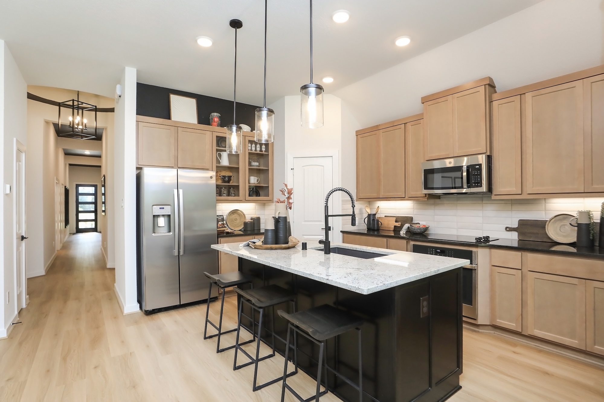 Modern kitchen with light wood cabinets, stainless steel appliances, a center island with a white countertop, black stools, pendant lights, and light wood flooring; a hallway is visible in the background.