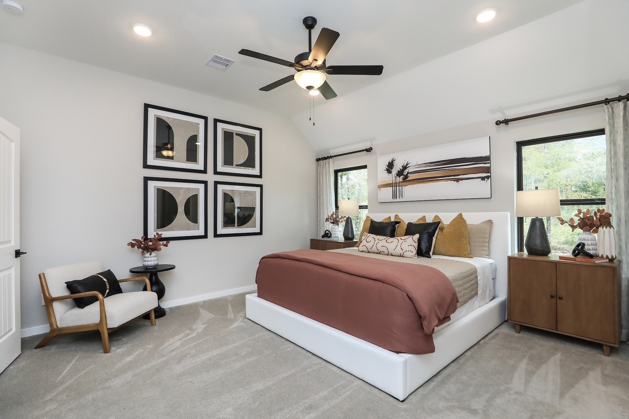 A modern bedroom with a white bed frame, rust-colored bedding, and decorative pillows. The room features abstract wall art, a ceiling fan, a wooden nightstand with lamps, a chair, and large windows with drapes.