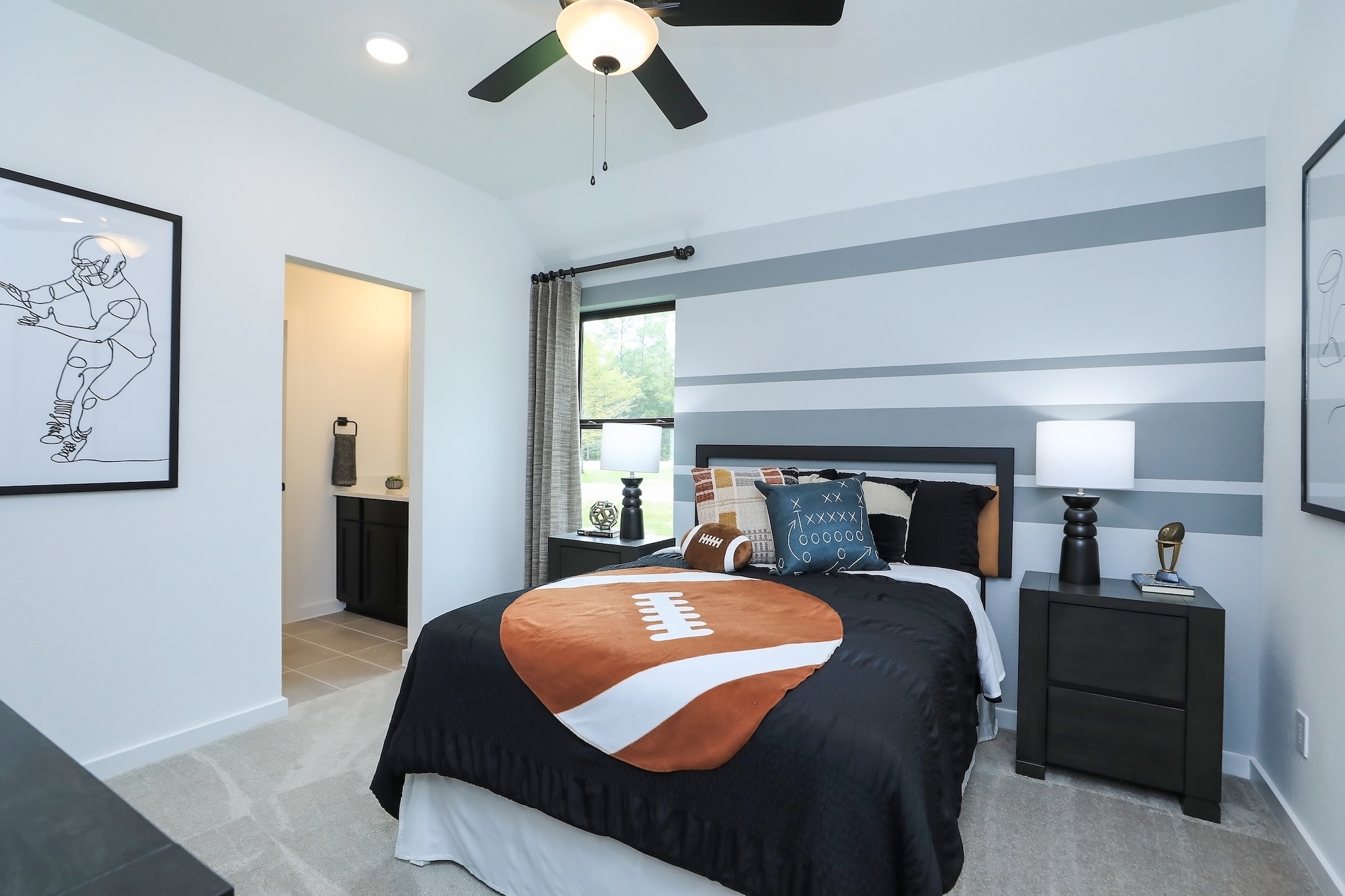 A modern bedroom with a football-themed bedspread, black and white decor, striped accent wall, two nightstands with lamps, framed sports art, and a window with gray curtains letting in natural light.