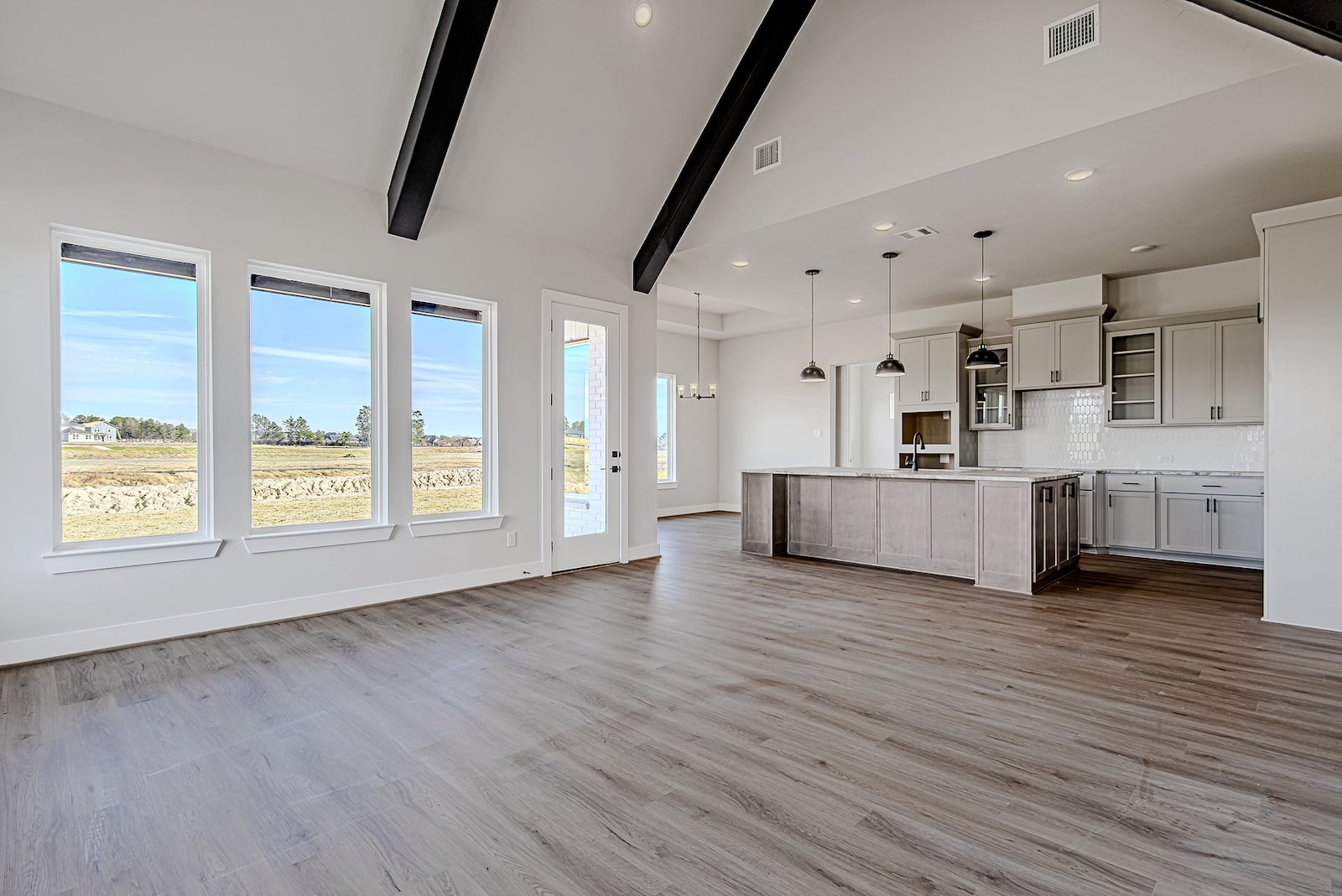 Modern, unfurnished open-concept living space with large windows, vaulted ceiling with exposed beams, wood-style flooring, and a kitchen featuring pendant lights and light-colored cabinets. Bright and spacious atmosphere.