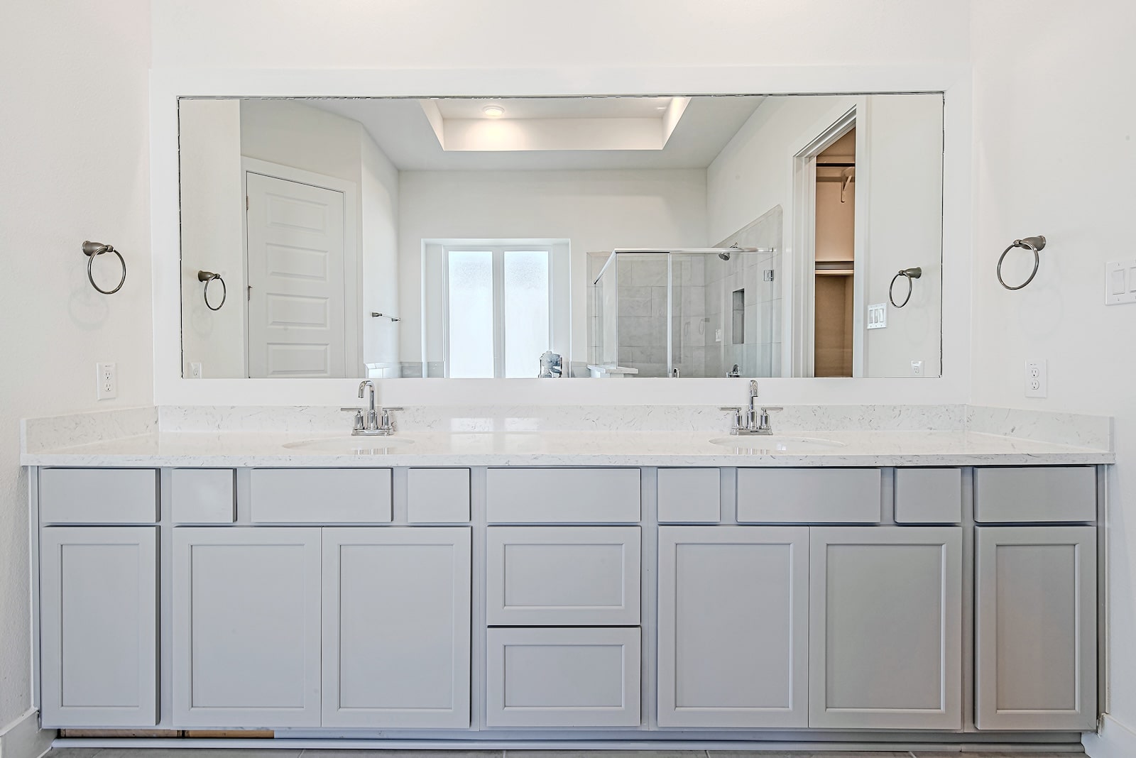 A modern bathroom with a large double-sink vanity, light gray cabinets, white countertops, two faucets, a wide mirror, and a walk-in shower visible in the background.