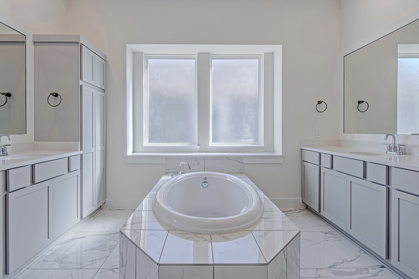 Spacious, modern bathroom with a central white oval bathtub, marble floors, large frosted windows, double sinks with mirrors, and white cabinetry on both sides.