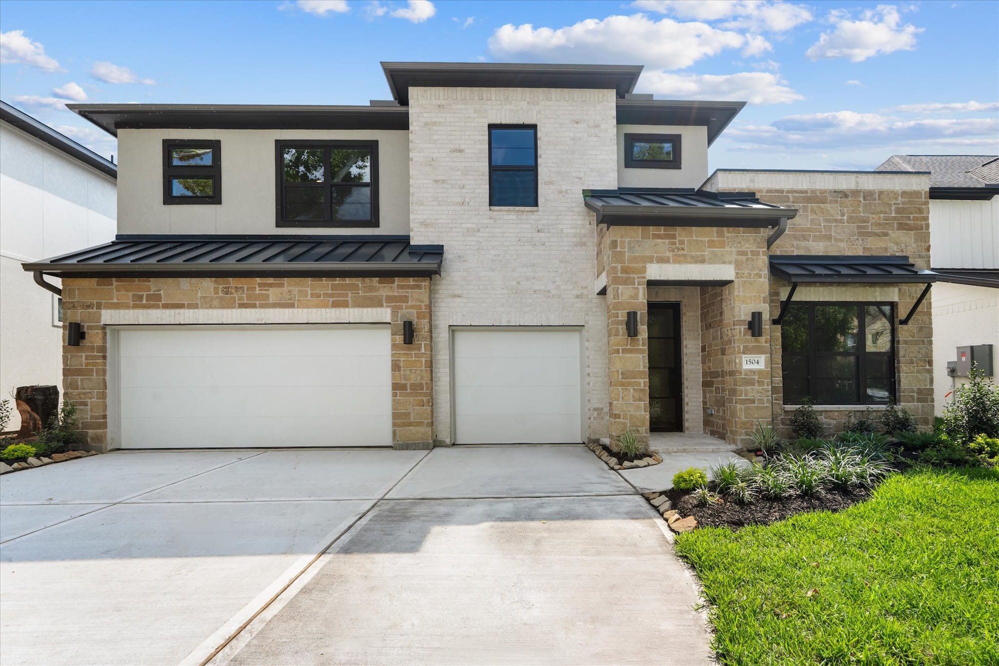 Modern two-story house with light brick and stone exterior, black trim, large windows, double garage, landscaped front yard, and a concrete driveway under a blue sky with scattered clouds.