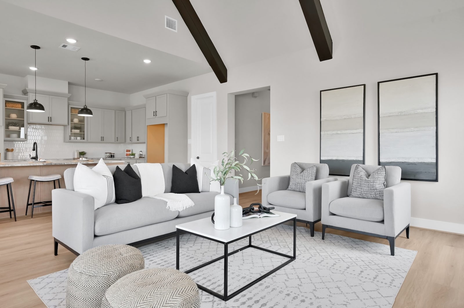 Modern living room with a light gray sofa, two armchairs, black and white pillows, a white coffee table, and minimalist decor. Kitchen with bar stools and pendant lights visible in the background.