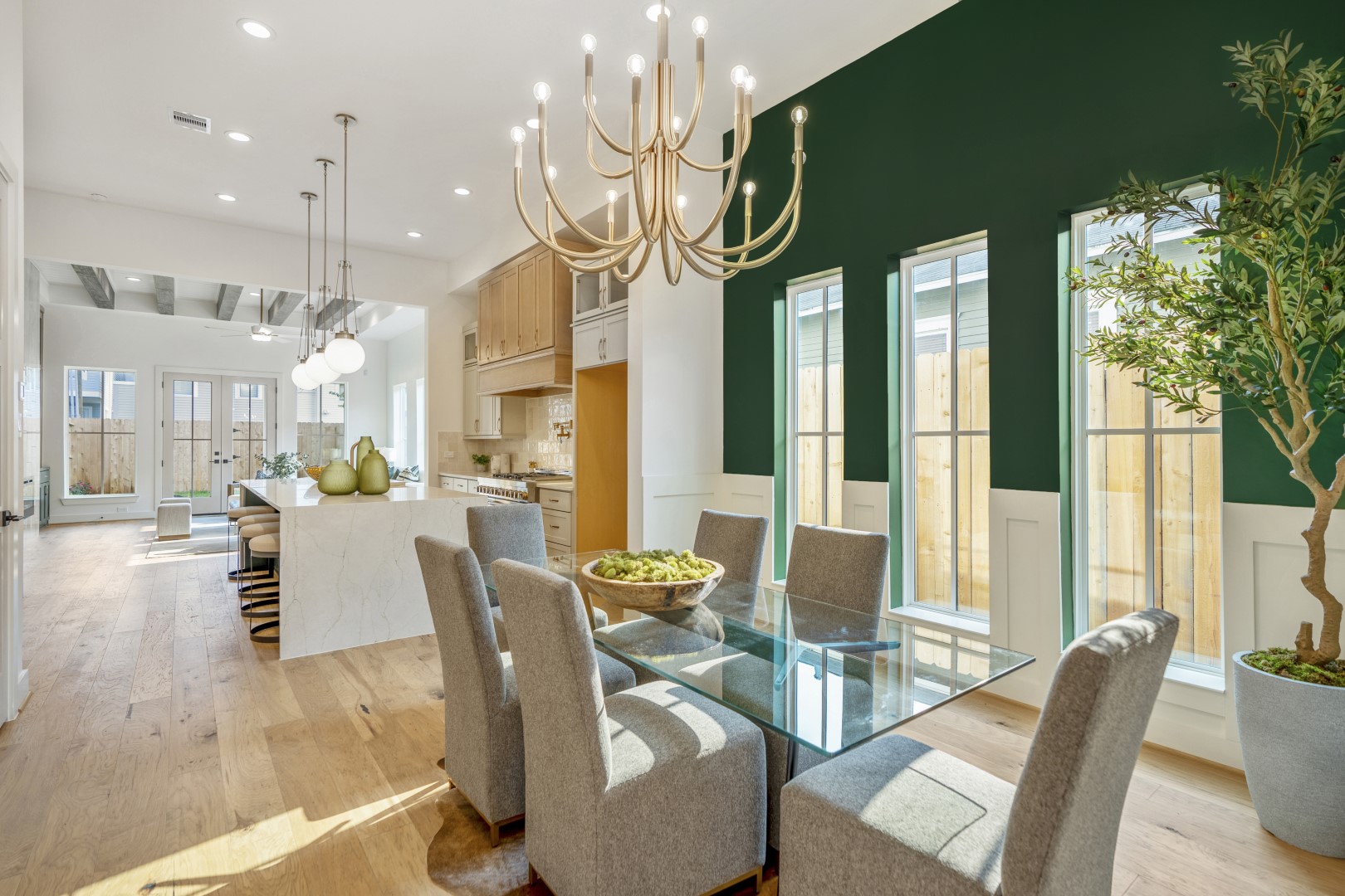 Modern dining room with a glass table, eight gray upholstered chairs, a gold chandelier, and a green accent wall with tall windows. The open layout connects to a bright kitchen with light wood cabinets and island seating.