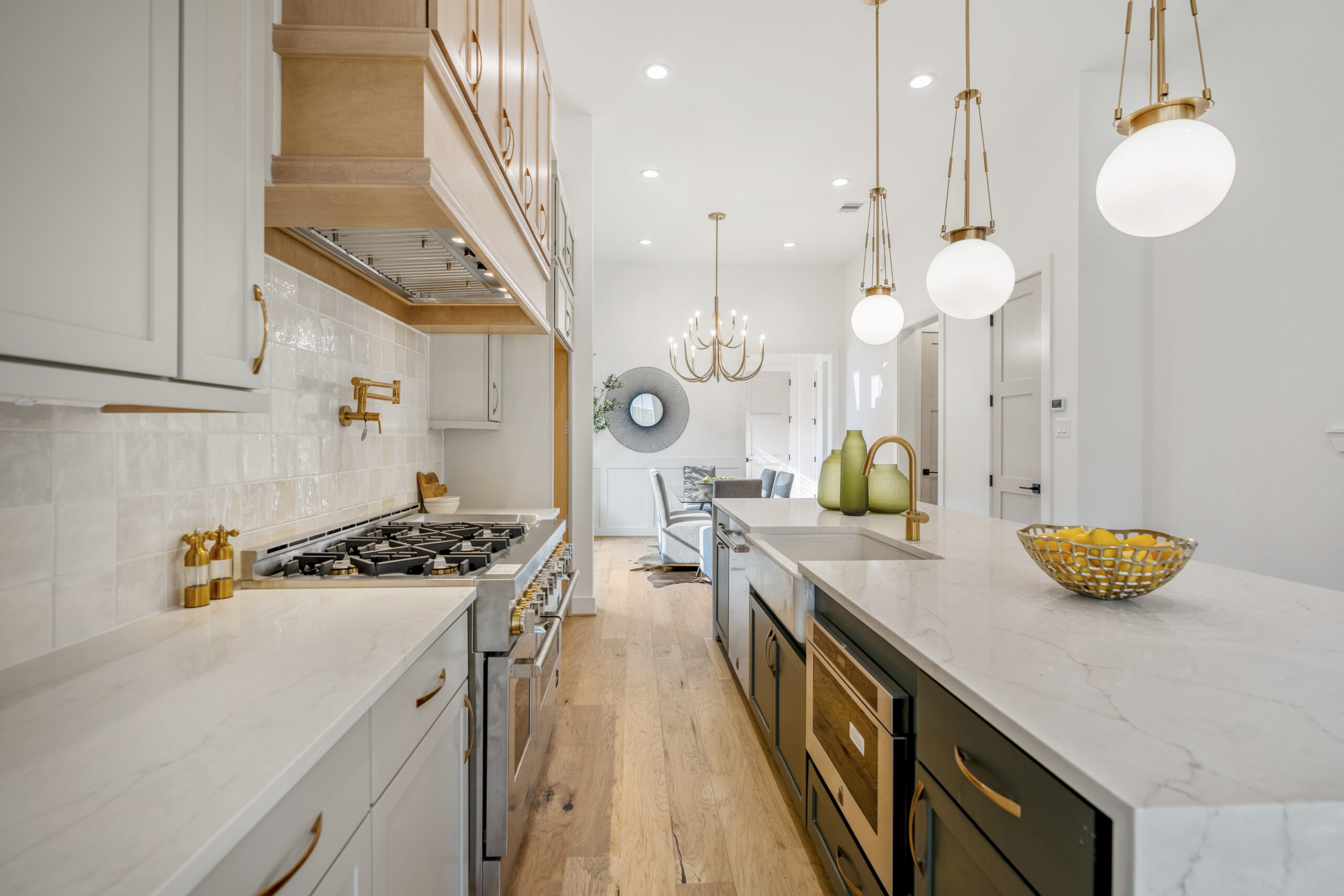 Modern kitchen with marble countertops, gold fixtures, and pendant lights. There is a gas stove, kitchen island with a bowl of lemons, and light wood flooring. In the background, a living area with a circular mirror is visible.