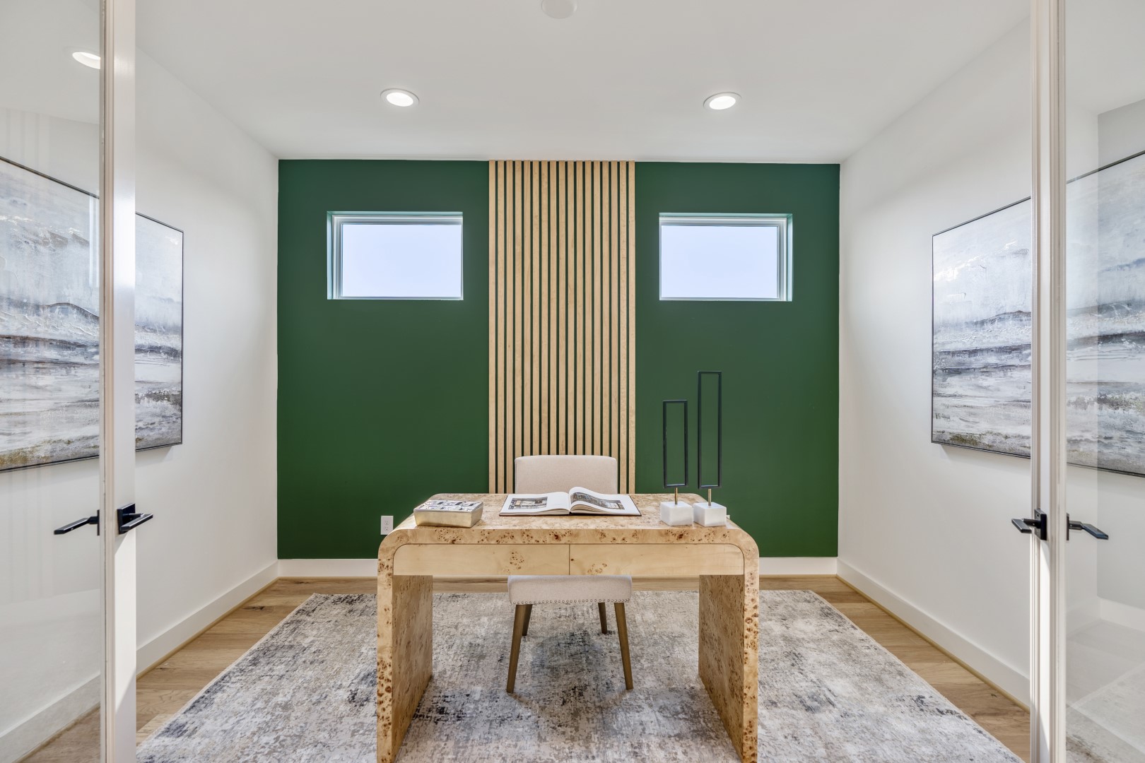 A modern home office with a wooden desk, beige chair, green accent wall with vertical wood slats, two small windows, abstract wall art, and a gray rug on light wood flooring.