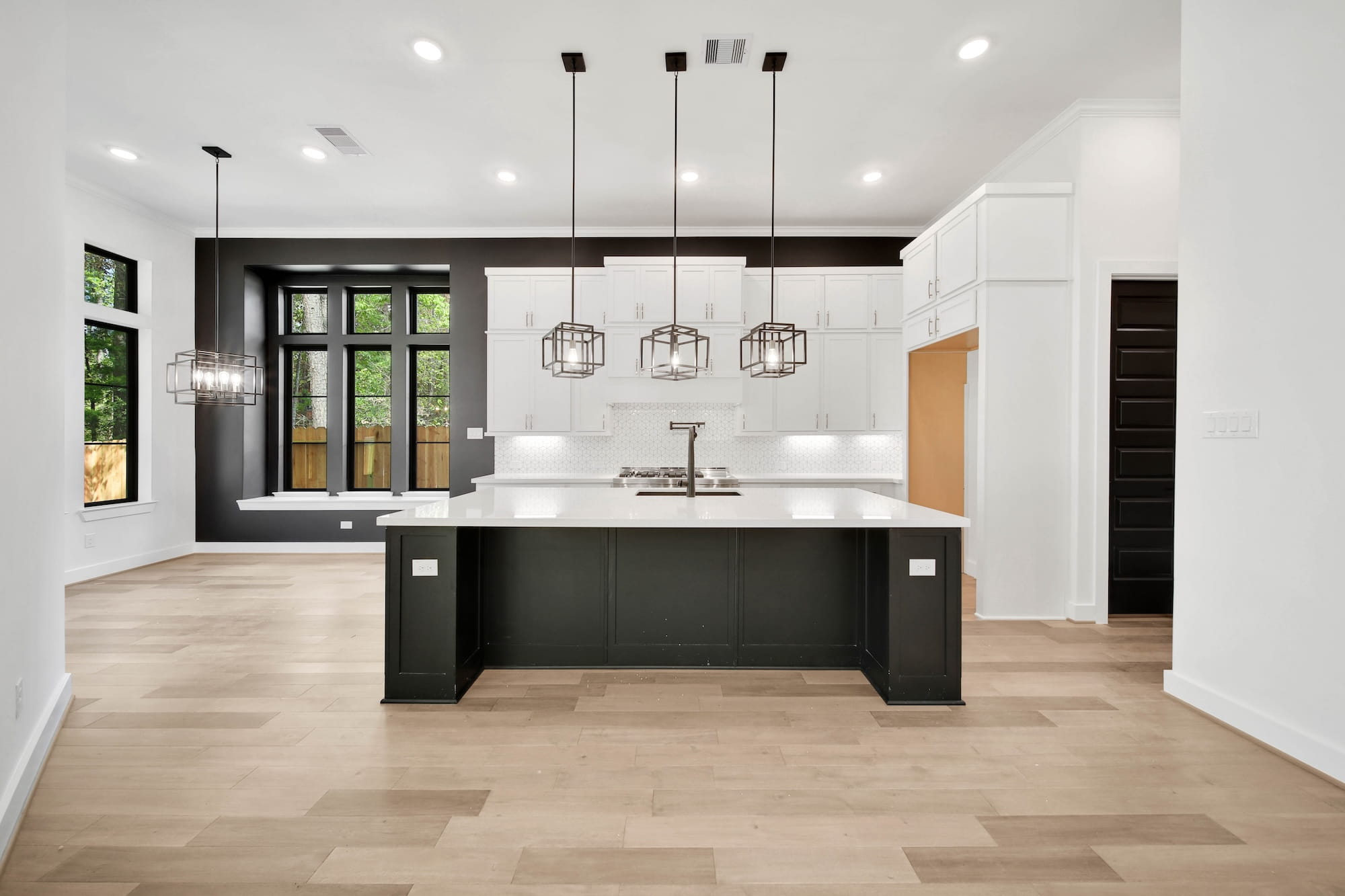 Modern kitchen with black and white color scheme, featuring a large island with pendant lights, white cabinets, built-in window seating, and light wood floors. Large windows provide natural light.