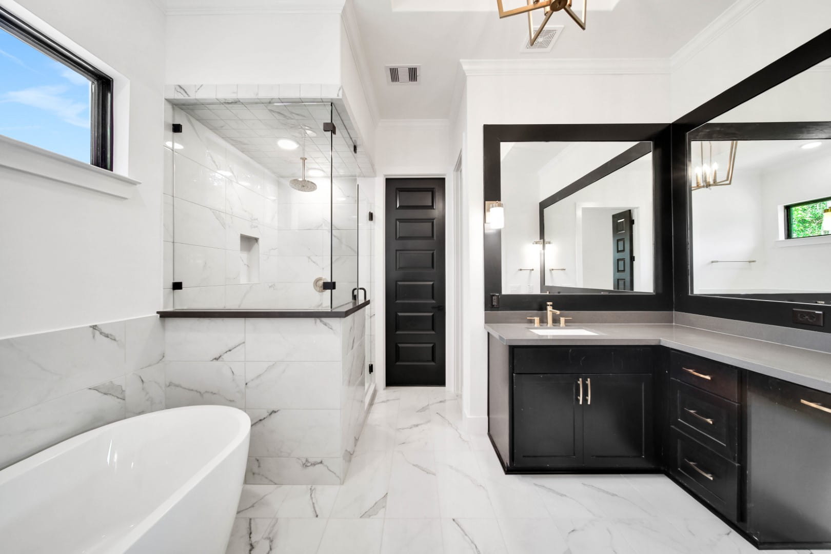 Modern bathroom with white marble floors and walls, a glass-enclosed shower, a freestanding bathtub, a large mirror above a dark vanity with gold fixtures, and a black door.