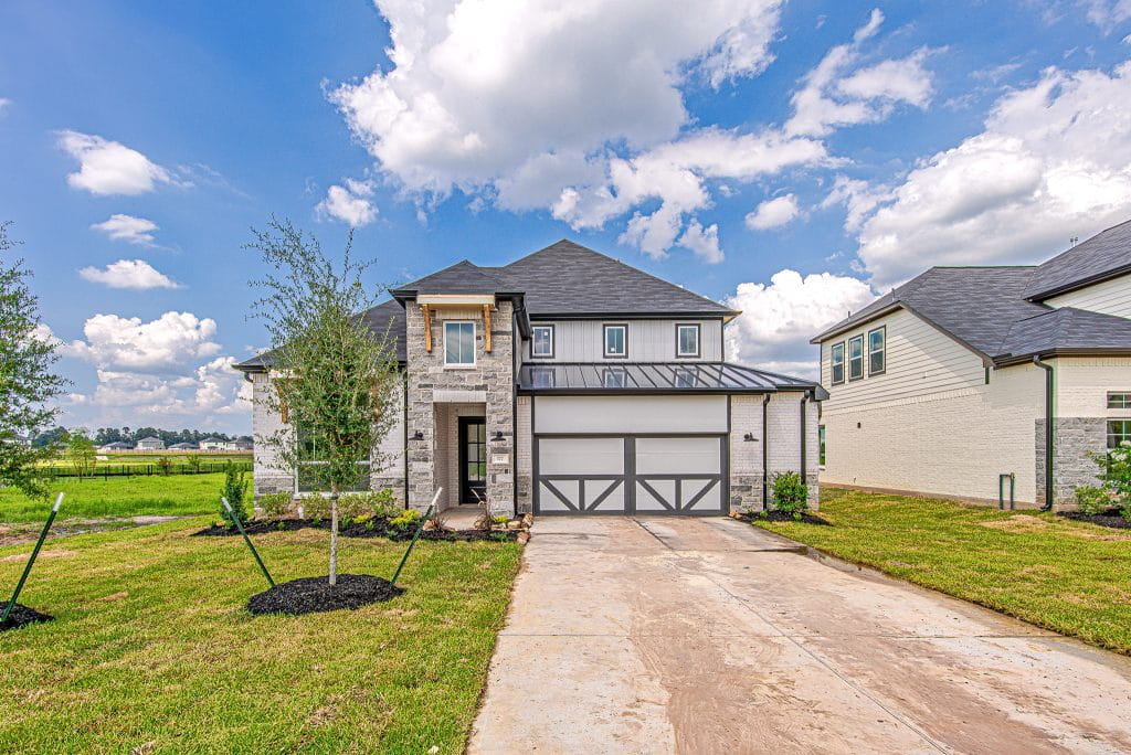 A modern two-story house with gray stone and white siding, a dark metal roof, and a two-car garage sits on a freshly landscaped lawn under a blue sky with scattered clouds.