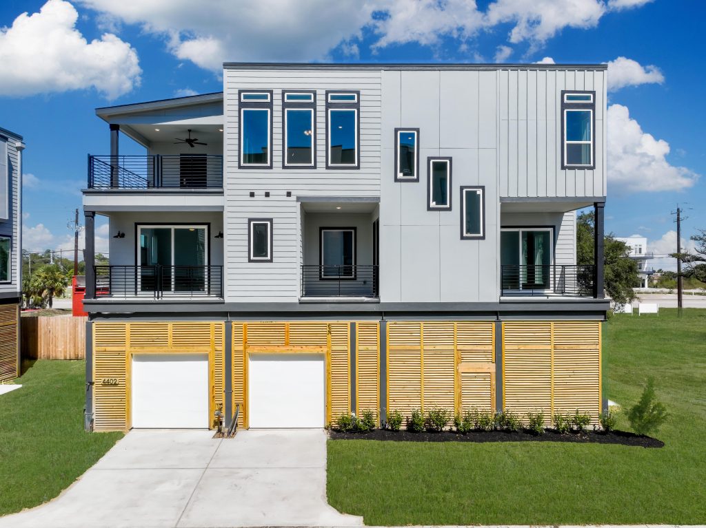 Modern three-story house with gray siding, large windows, balconies with black railings, and a double garage. The front yard has green grass and a small garden along the wooden slats on the ground floor.